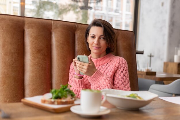 Una donna in un ristorante con un maglione caldo sta guardando qualcosa al telefono, una deliziosa colazione sana e matcha latte sul tavolo