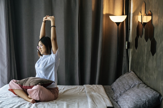 Una donna in una camicia bianca seduta sul letto e sollevando entrambe le braccia.