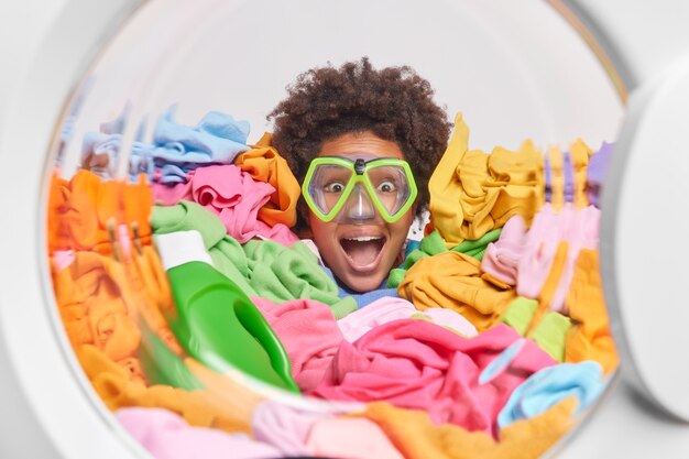 Una donna positiva con i capelli ricci indossa una maschera da snorkeling bloccata in posa per il bucato al tamburo della lavatrice essendo molto sorpresa da qualcosa