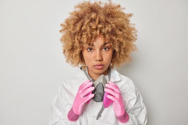 Una donna seria e attenta con i capelli ricci tiene le mani sulla maschera antigas indossa tuta protettiva e maschera antigas guarda direttamente la fotocamera isolata su sfondo bianco Concetto di inquinamento di pericolo chimico di persone