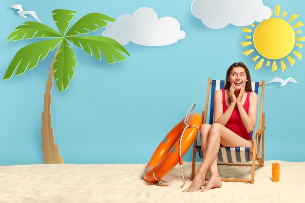 Una donna sognante e positiva gode di una calda giornata sulla costa, si siede sulla sdraio, indossa un bikini rosso, usa una crema solare per proteggere la pelle dal sole