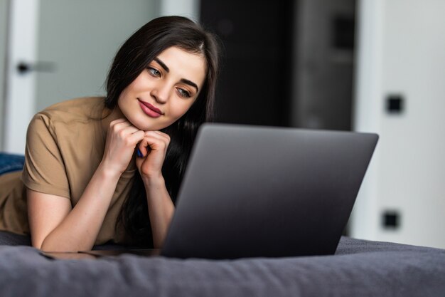 Una donna sorridente sdraiata sul letto davanti al suo laptop. Lavorare da casa in quarantena. Distanziamento sociale Autoisolamento