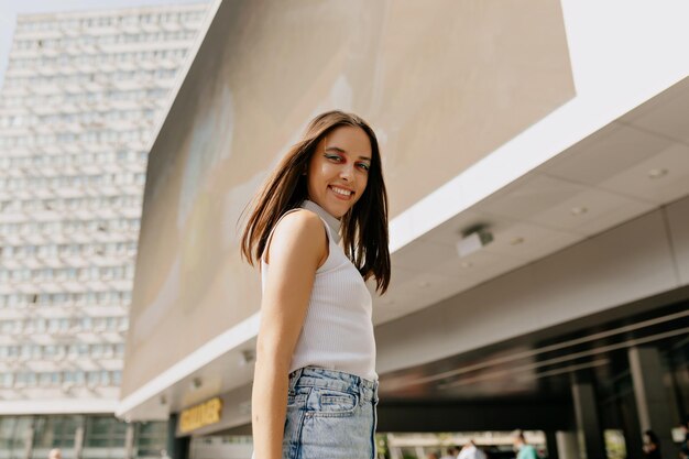 Una donna spettacolare alla moda con i capelli scuri che indossa una maglietta bianca e jeans è in posa davanti alla telecamera all'aperto alla luce del sole nel centro della città