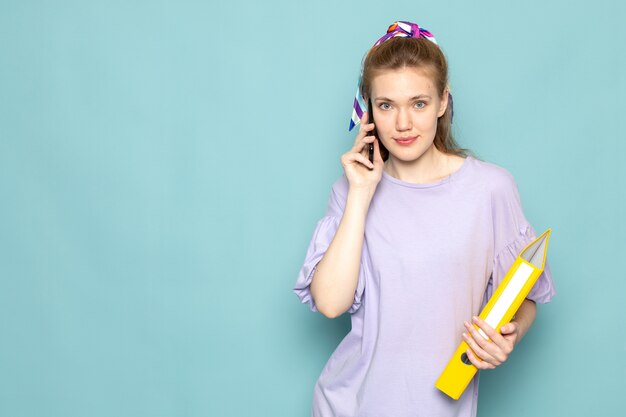 Una femmina attraente di vista frontale in camicia-vestito blu che tiene archivi gialli e che parla sul telefono sull'azzurro