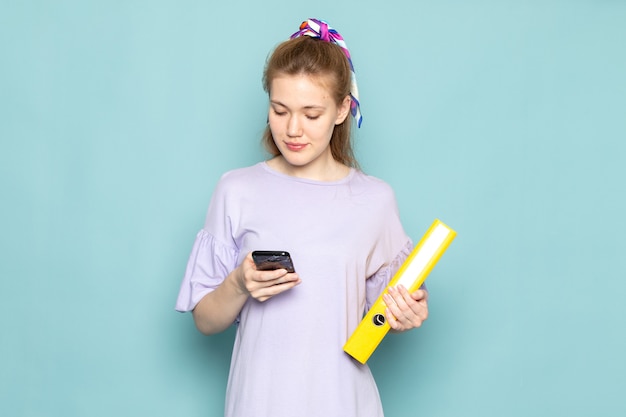 Una femmina attraente di vista frontale in camicia-vestito blu che tiene archivi gialli e usando un telefono sull'azzurro