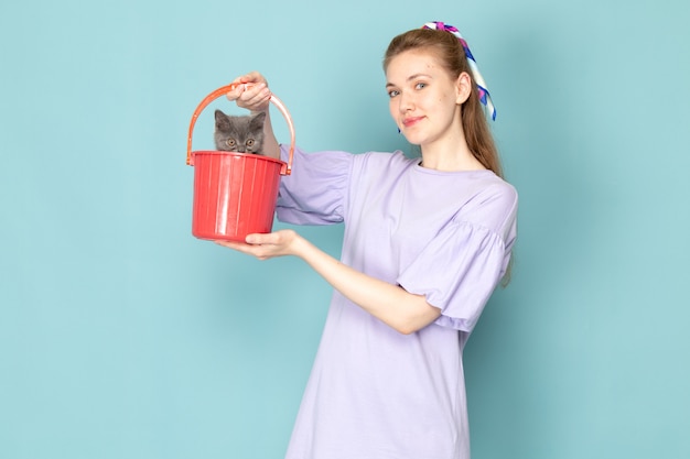 Una femmina attraente di vista frontale in camicia viola che tiene secchio rosso con il gattino sveglio sull'azzurro