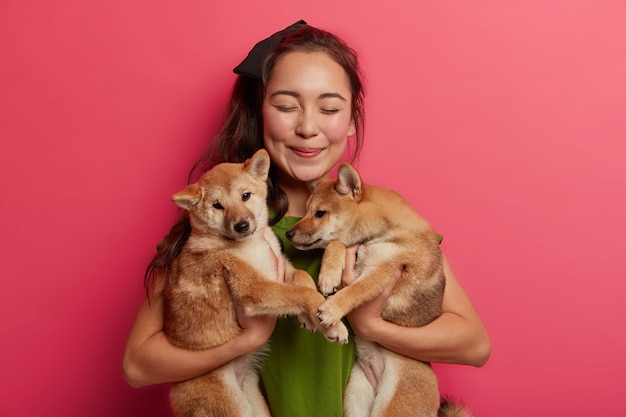 Una fortunata donna orientale ha trovato due cuccioli di razza per strada, trova un ospite per i cani Shiba Inu, essendo amante degli animali domestici, è felice con gli animali su sfondo rosa.