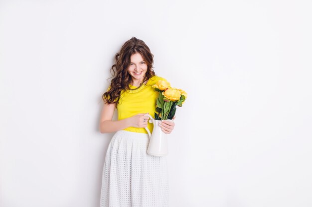 Una foto in studio di una ragazza in piedi e con in mano un vaso bianco con fiori. La ragazza ha lunghi capelli castani ondulati e indossa una maglietta gialla e una gonna bianca. La ragazza sorride ampiamente.