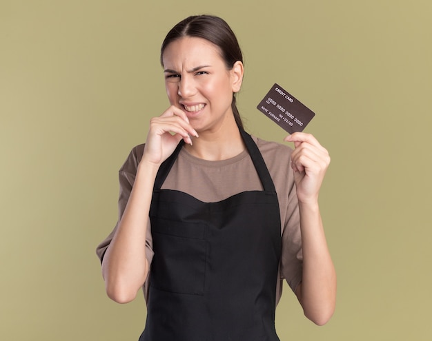 Una giovane barbiere bruna scontenta in uniforme mette la mano sul mento e tiene la carta di credito