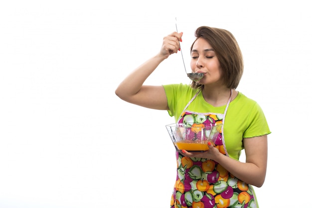 Una giovane bella casalinga di vista frontale in mantello variopinto della camicia verde che mescola le uova liquide che preparano il pasto che prova sulla cucina bianca della femmina della casa del fondo