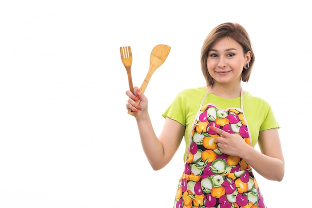 Una giovane bella casalinga di vista frontale nel capo variopinto della camicia verde che sorride tenendo l'elettrodomestico da cucina di legno sulla cucina bianca di pulizia della casa del fondo