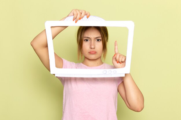 Una giovane bella ragazza di vista frontale in maglietta rosa che posa con l'espressione scontenta di carta a forma di TV bianca sul verde