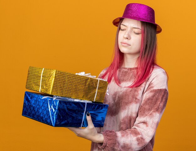 Una giovane bella ragazza scontenta che indossa un cappello da festa che tiene in mano e guarda le scatole regalo