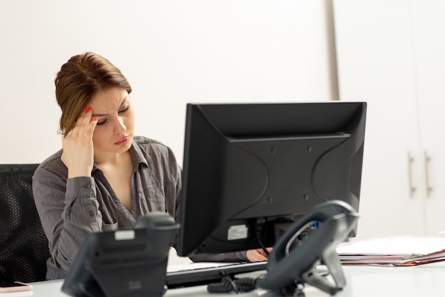 Una giovane bella signora di vista frontale in camicia grigia che per mezzo del suo pc che si siede dentro il suo ufficio che pensa calcolando durante l'attività di lavoro di costruzione di giorno