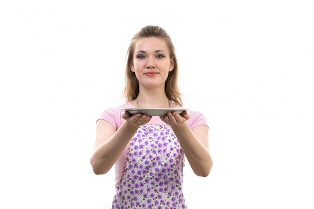 Una giovane casalinga attraente di vista frontale in capo variopinto della camicia rosa che tiene posa sorridente del piatto bianco sulla femmina bianca della cucina di cucina del fondo