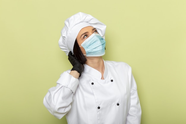 Una giovane cuoca femminile di vista frontale in vestito bianco da cuoco e berretto che indossa guanti e maschera sterile in posa sul colore dell'alimento del lavoro della signora della parete verde