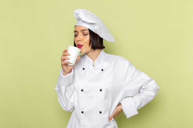 Una giovane cuoca femminile di vista frontale in vestito bianco del cuoco e cappuccio che tiene il bicchiere di plastica sul colore della cucina dell'alimento del lavoro della signora della parete verde