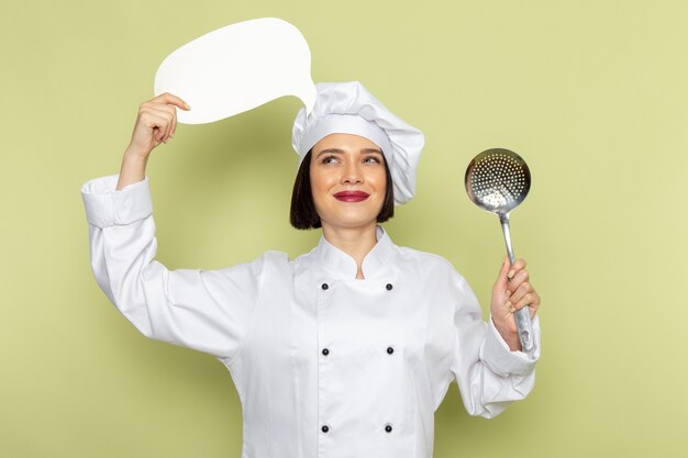 Una giovane cuoca femminile di vista frontale in vestito bianco del cuoco e cappuccio che tiene il cucchiaio e segno bianco sul colore della cucina dell'alimento del lavoro della signora della parete verde