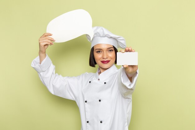 Una giovane cuoca femminile di vista frontale in vestito bianco e cappuccio del cuoco che tiene segno bianco sul colore verde della cucina dell'alimento della signora del lavoro della parete