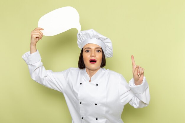 Una giovane cuoca femminile di vista frontale in vestito bianco e cappuccio del cuoco che tiene segno bianco sul colore verde della cucina dell'alimento della signora del lavoro della parete