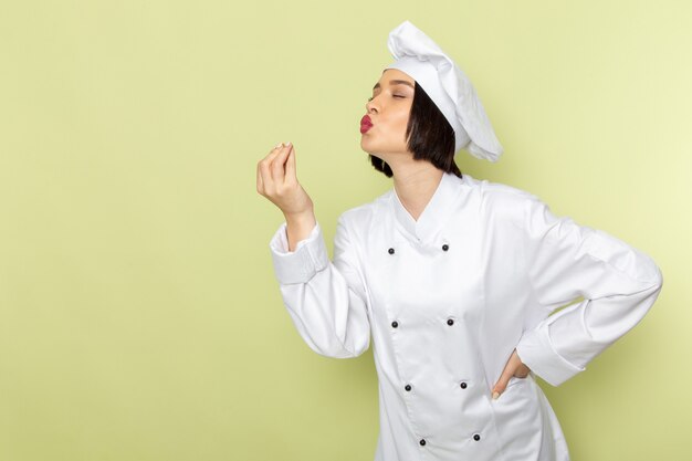 Una giovane cuoca femminile di vista frontale in vestito e cappuccio bianchi del cuoco che posano e che mostrano il gesto saporito sul colore della cucina dell'alimento del lavoro della signora della parete verde