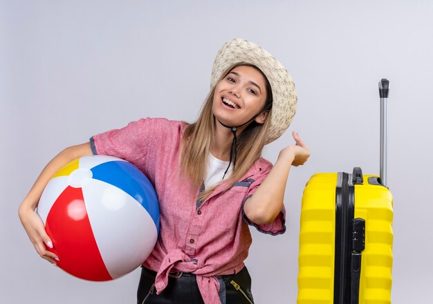 Una giovane donna abbastanza adorabile che indossa la camicia rossa e il cappello da sole che sorride mentre tiene la sfera gonfiabile su una parete bianca