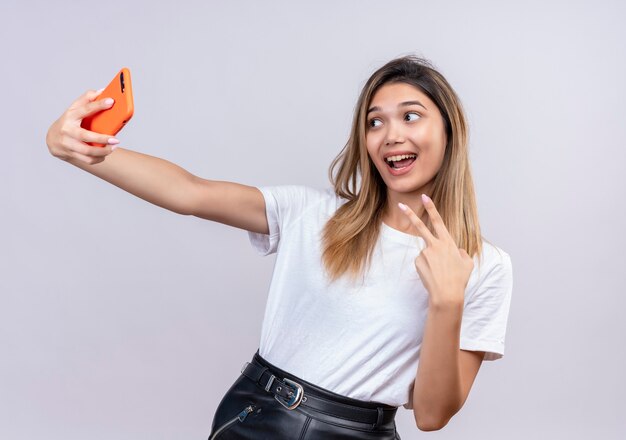 Una giovane donna adorabile felice in maglietta bianca che parla sulla videochiamata utilizzando smartphone e che mostra un gesto di due mani su un muro bianco