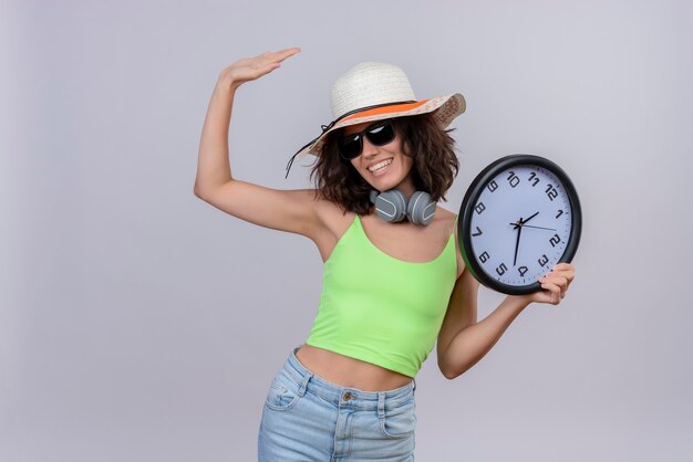 Una giovane donna allegra con i capelli corti in top corto verde che indossa occhiali da sole e cappello da sole in possesso di un orologio da parete e alzando la mano su uno sfondo bianco