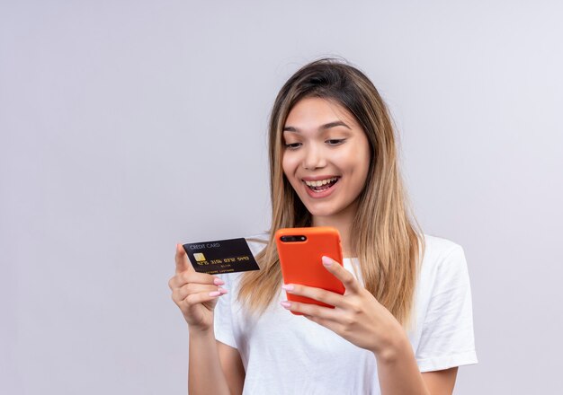 Una giovane donna allegra in camicia bianca che digita sul telefono cellulare mentre si tiene la carta di credito su un muro bianco