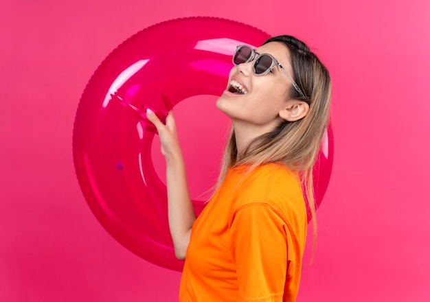 Una giovane donna allegra in una maglietta arancione che indossa occhiali da sole guardando il lato mentre tiene l'anello gonfiabile rosa su una parete rosa