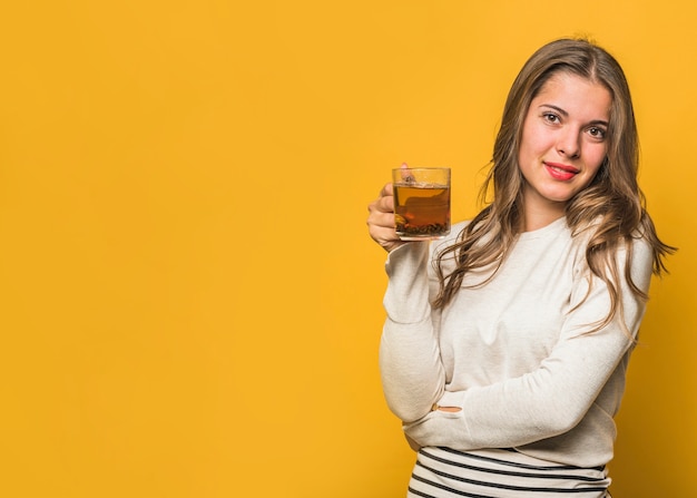 Una giovane donna attraente che tiene tazza di tisana in mano in piedi contro il contesto giallo