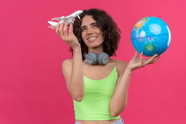 Una giovane donna attraente felice con i capelli corti nella parte superiore del raccolto verde in cuffie che tengono il globo e che esaminano l'aereo del giocattolo