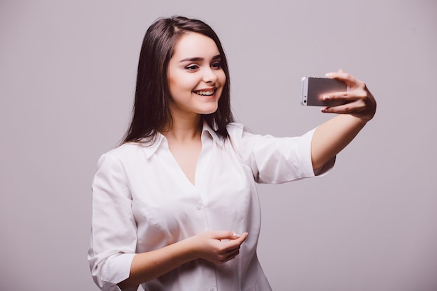 Una giovane donna attraente sorridente che tiene una macchina fotografica digitale con la sua mano e che prende un autoritratto del selfie, isolato su fondo bianco.