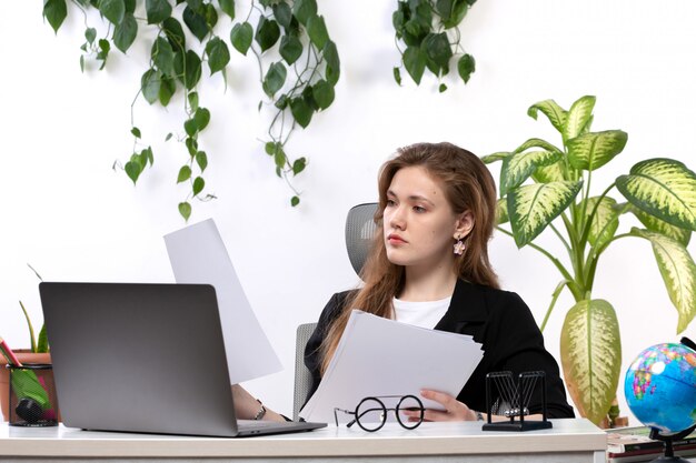 Una giovane donna bella vista frontale in camicia bianca e giacca nera, lavorando con documenti usando il suo computer portatile davanti al tavolo con foglie appese