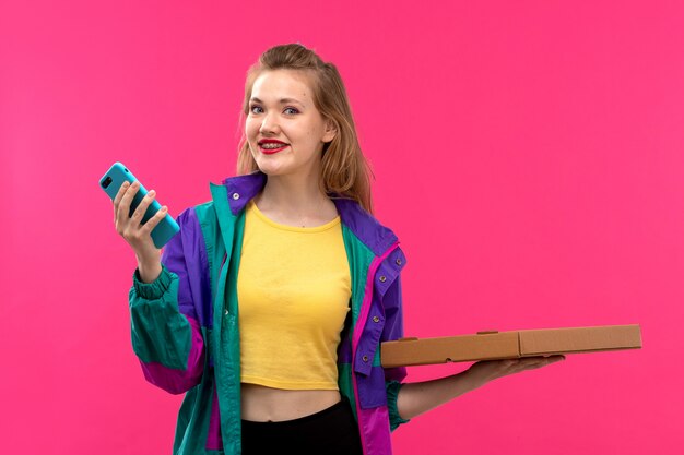 Una giovane donna bella vista frontale in giacca colorata arancione camicia nera pantaloni sorridenti utilizzando il suo telefono tenendo la pizza