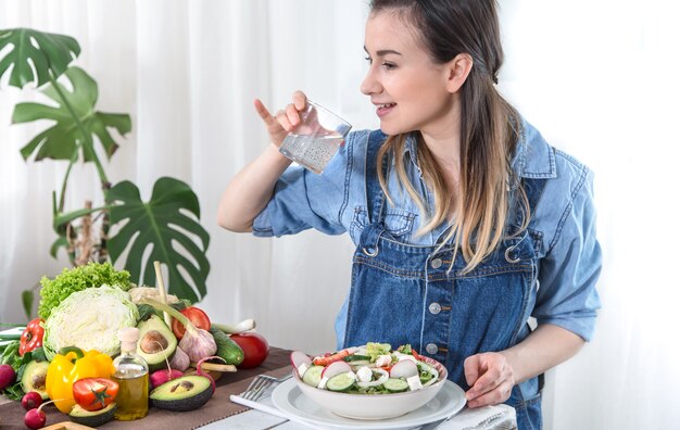 Una giovane donna beve acqua a un tavolo con verdure su sfondo chiaro, vestita con abiti in denim. Concetto di cibo e bevande sani.