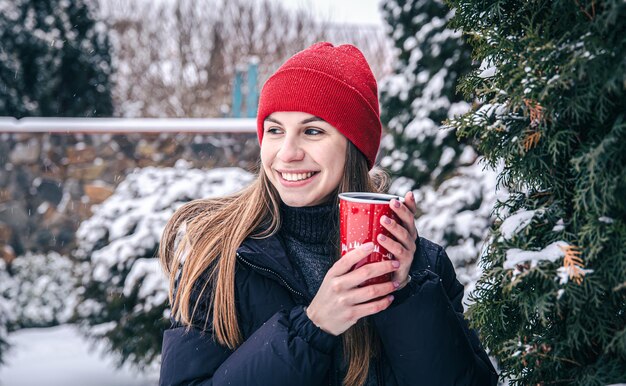 Una giovane donna beve una bevanda calda da una tazza termica rossa in inverno