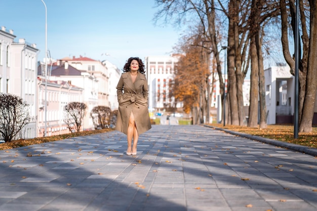 una giovane donna cammina per la città autunnale, una femmina dal corpo felice con i capelli ricci in un impermeabile.