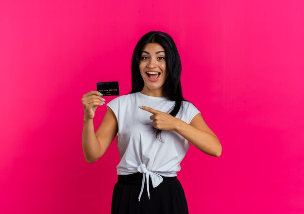 Una giovane donna caucasica eccitata tiene e indica la carta di credito guardando la telecamera