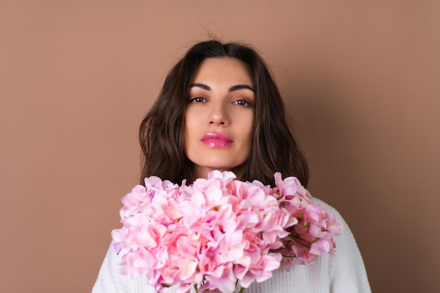 Una giovane donna con capelli ondulati e voluminosi su uno sfondo beige con un rossetto rosa brillante e un lucidalabbra in un maglione bianco tiene un mazzo di fiori rosa