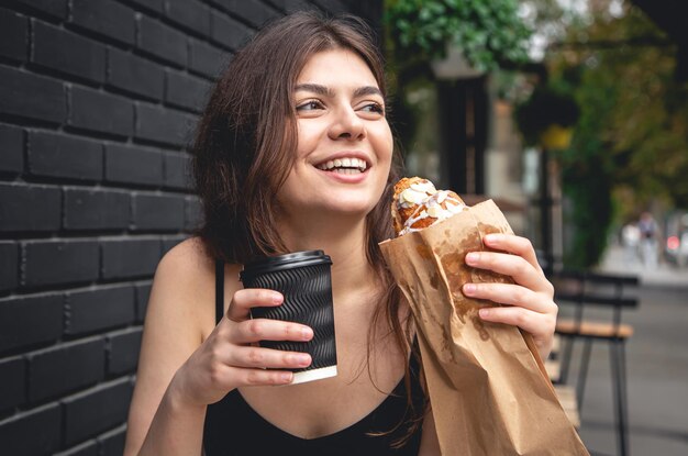 Una giovane donna con un croissant e una tazza di caffè contro un muro di mattoni neri