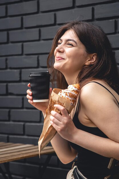 Una giovane donna con un croissant e una tazza di caffè contro un muro di mattoni neri