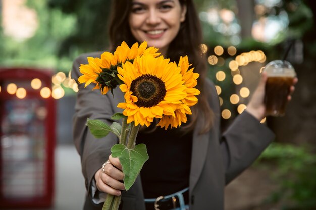 Una giovane donna con un mazzo di girasoli e una bevanda fredda al caffè in città