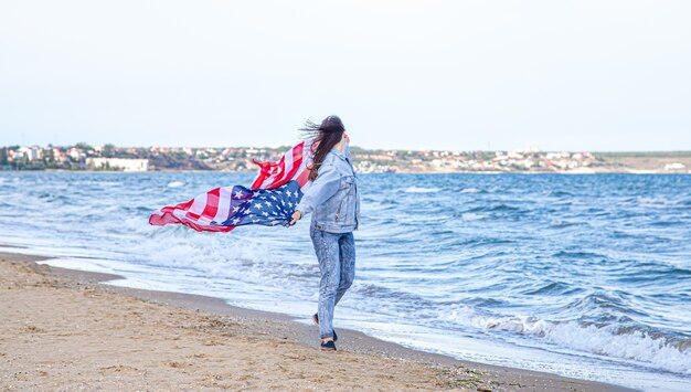 Una giovane donna con una bandiera americana corre in riva al mare. Il concetto di patriottismo e celebrazioni del giorno dell'indipendenza.