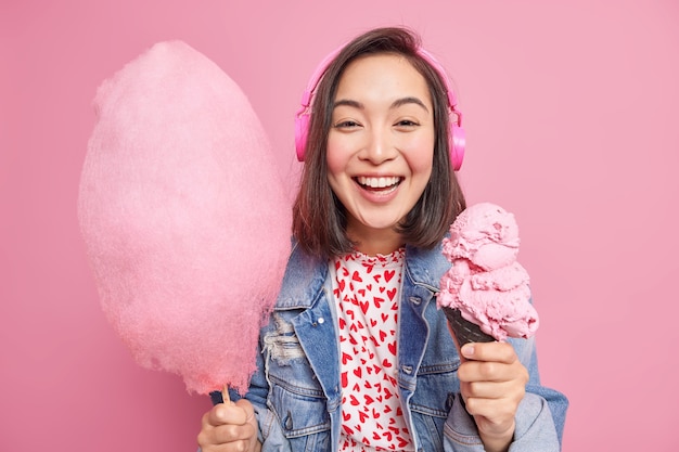 Una giovane donna dai capelli abbastanza scura con espressione positiva tiene gelato congelato e zucchero filato ascolta la traccia audio tramite cuffie vestita con abiti alla moda isolati sul muro rosa