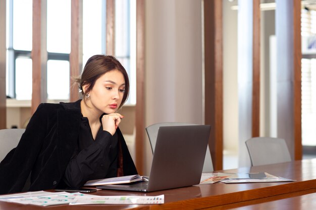 Una giovane donna di affari attraente di vista frontale in rivestimento nero della camicia nera facendo uso del suo computer portatile d'argento che lavora dentro la sua costruzione di lavoro del lavoro d'ufficio