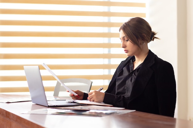 Una giovane donna di affari attraente di vista frontale in rivestimento nero della camicia nera facendo uso della sua lettura d'argento di scrittura del computer portatile che funziona dentro la sua costruzione di lavoro del lavoro d'ufficio