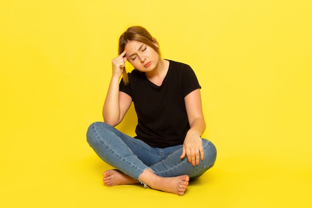 Una giovane donna di vista frontale stanca e depressa seduta in camicia nera e blue jeans su giallo