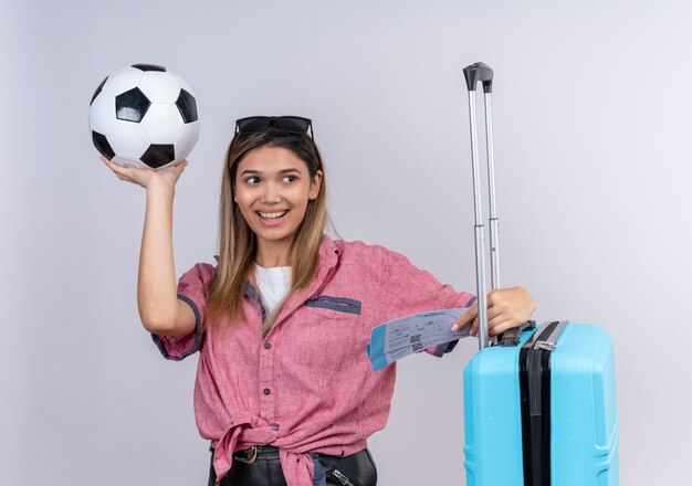 Una giovane donna felice che indossa la camicia rossa e gli occhiali da sole guardando il lato mentre si tiene la palla con i biglietti aerei e la valigia blu su un muro bianco