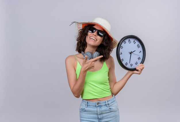 Una giovane donna felice con i capelli corti in top corto verde che indossa occhiali da sole e cappello da sole che punta a un orologio da parete su uno sfondo bianco